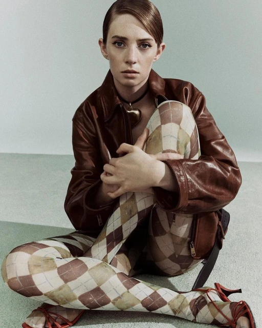 A woman with styled hair sits in a brown leather jacket and patterned tights, gazing calmly at the camera in a studio portrait.
