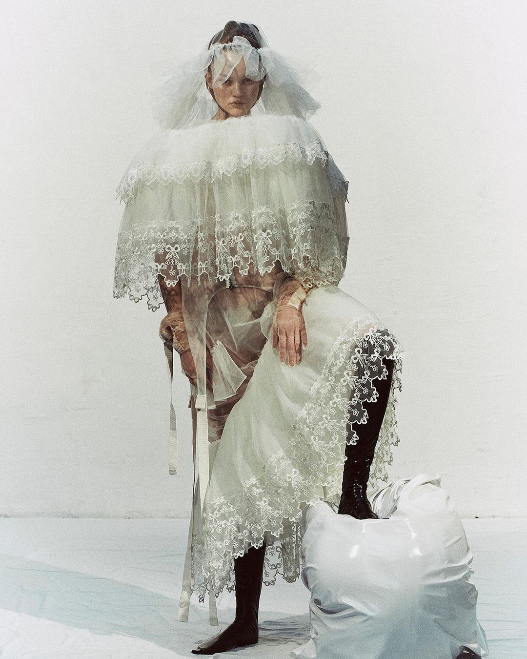 Fashionable woman in frilly white dress and veil poses in a studio setting. The dress features lace and frills and she has one foot on a white object.