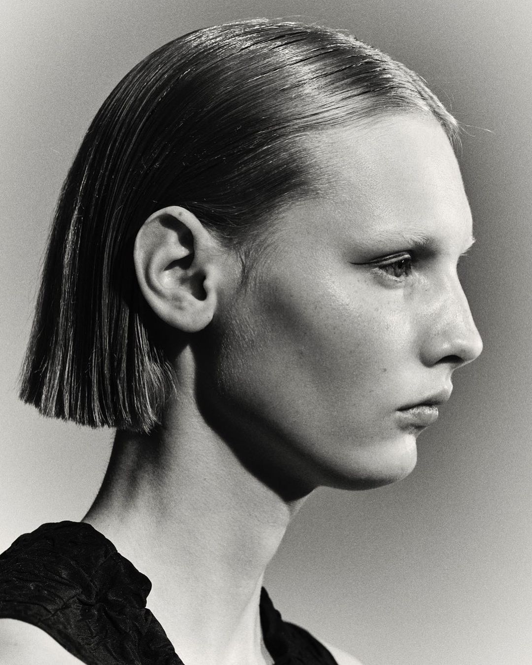 Profile portrait of a model with sleek short hair and dark top against a neutral gradient studio background.