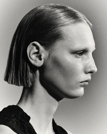 Profile portrait of a model with sleek short hair and dark top against a neutral gradient studio background.