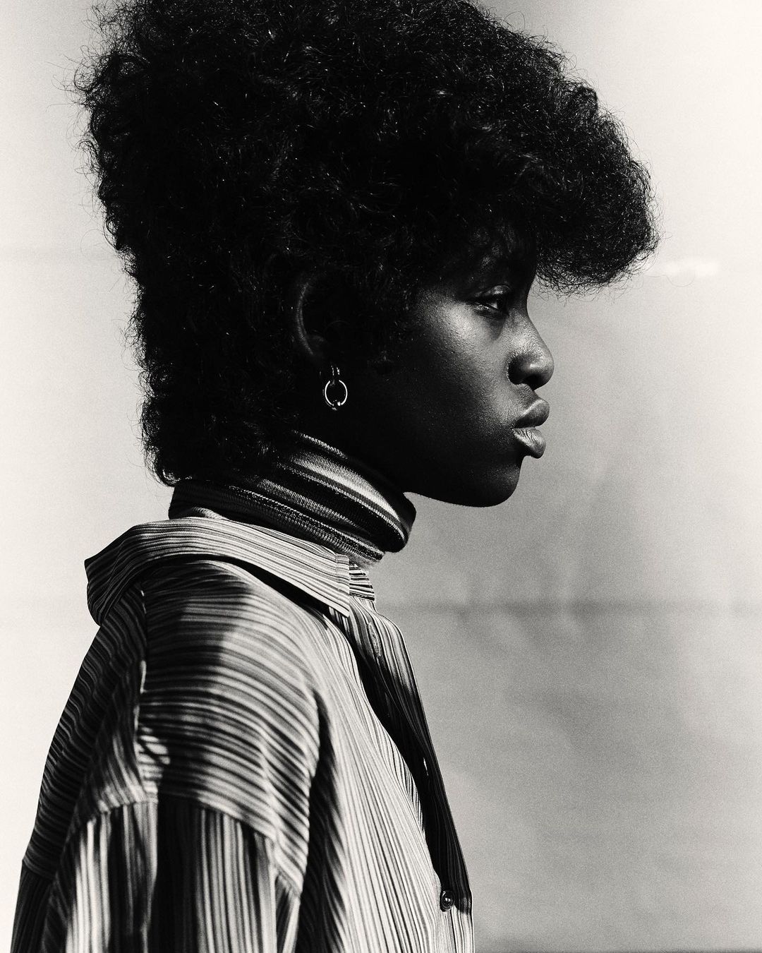 Black and white profile portrait of a woman with voluminous afro hair wearing a striped turtleneck sweater and collared shirt