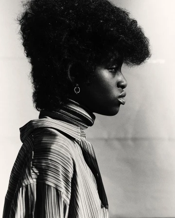 Black and white profile portrait of a woman with voluminous afro hair wearing a striped turtleneck sweater and collared shirt