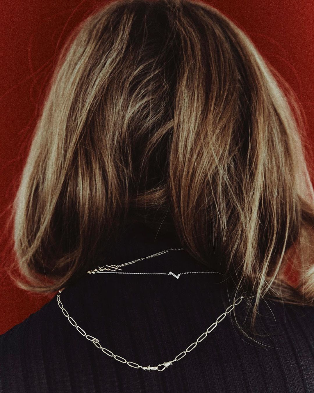 Blonde hair and silver chain necklaces on an adult female against a red background, shot from the back, fashion and beauty.