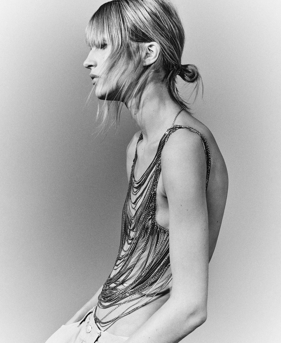 Black-and-white portrait of an adult woman with blonde hair, wearing a chainmail-style top, side profile against a plain background.