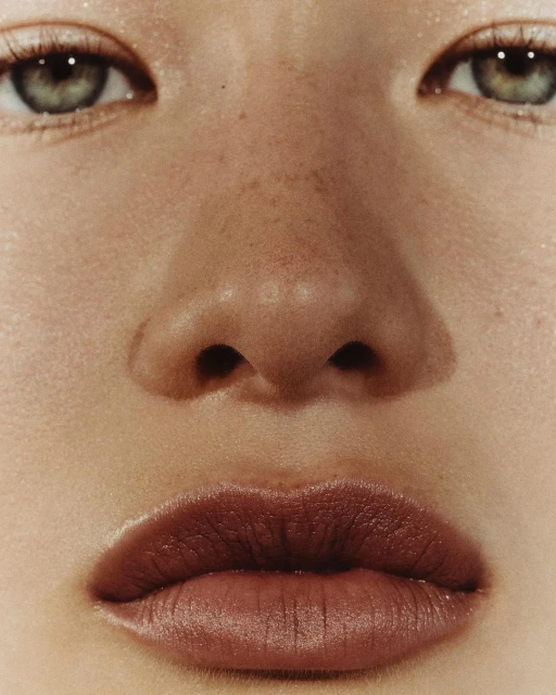 Close-up portrait of a young adult female face with fair skin, freckles, green eyes, and dark lipstick in a beauty shot.