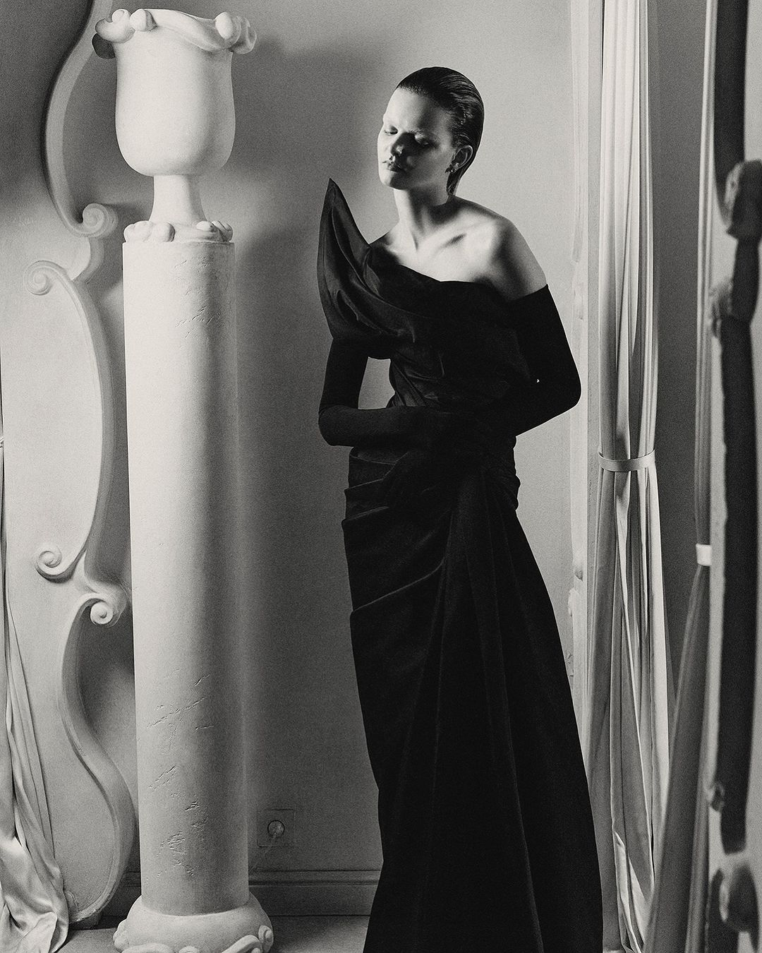 Black and white fashion shot of a woman in a dress with shoulder detail standing near architectural column and window light.