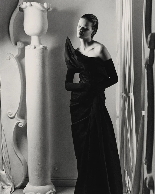 Black and white fashion shot of a woman in a dress with shoulder detail standing near architectural column and window light.