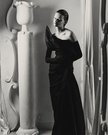Black and white fashion shot of a woman in a dress with shoulder detail standing near architectural column and window light.