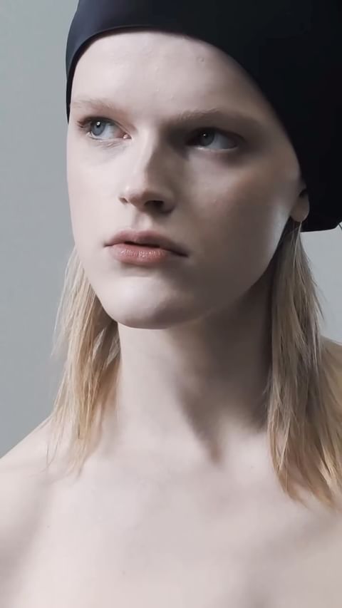 Close-up studio portrait of a light-skinned young adult female with blonde hair, wearing a black hat and light makeup, looking off-camera.