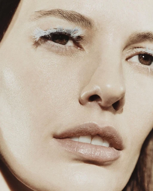Close-up beauty shot features a fair-skinned woman with creative white eyelashes, full pink lips, and fair skin with an indoor backdrop.