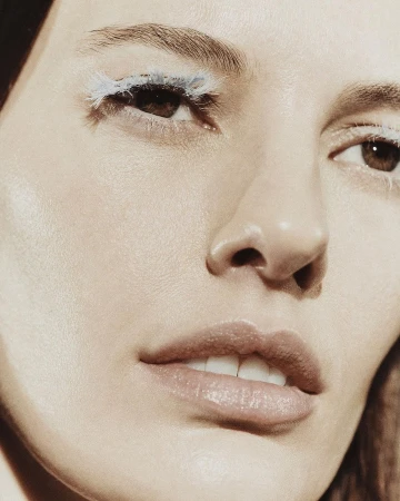 Close-up beauty shot features a fair-skinned woman with creative white eyelashes, full pink lips, and fair skin with an indoor backdrop.