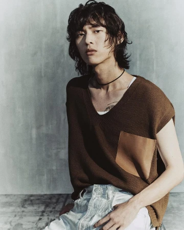 An Asian male fashion model poses in a sleeveless top and light-wash jeans against a textured wall in a portrait-style fashion shot.