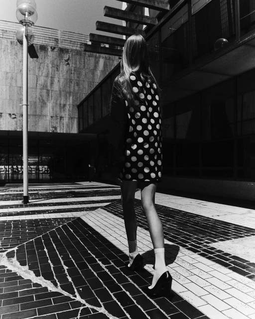 Woman in a polka dot dress walking on a tiled surface near a modern building, captured in a black and white outdoor shot.
