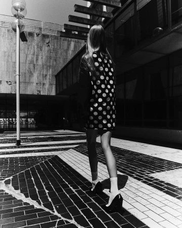 Woman in a polka dot dress walking on a tiled surface near a modern building, captured in a black and white outdoor shot.