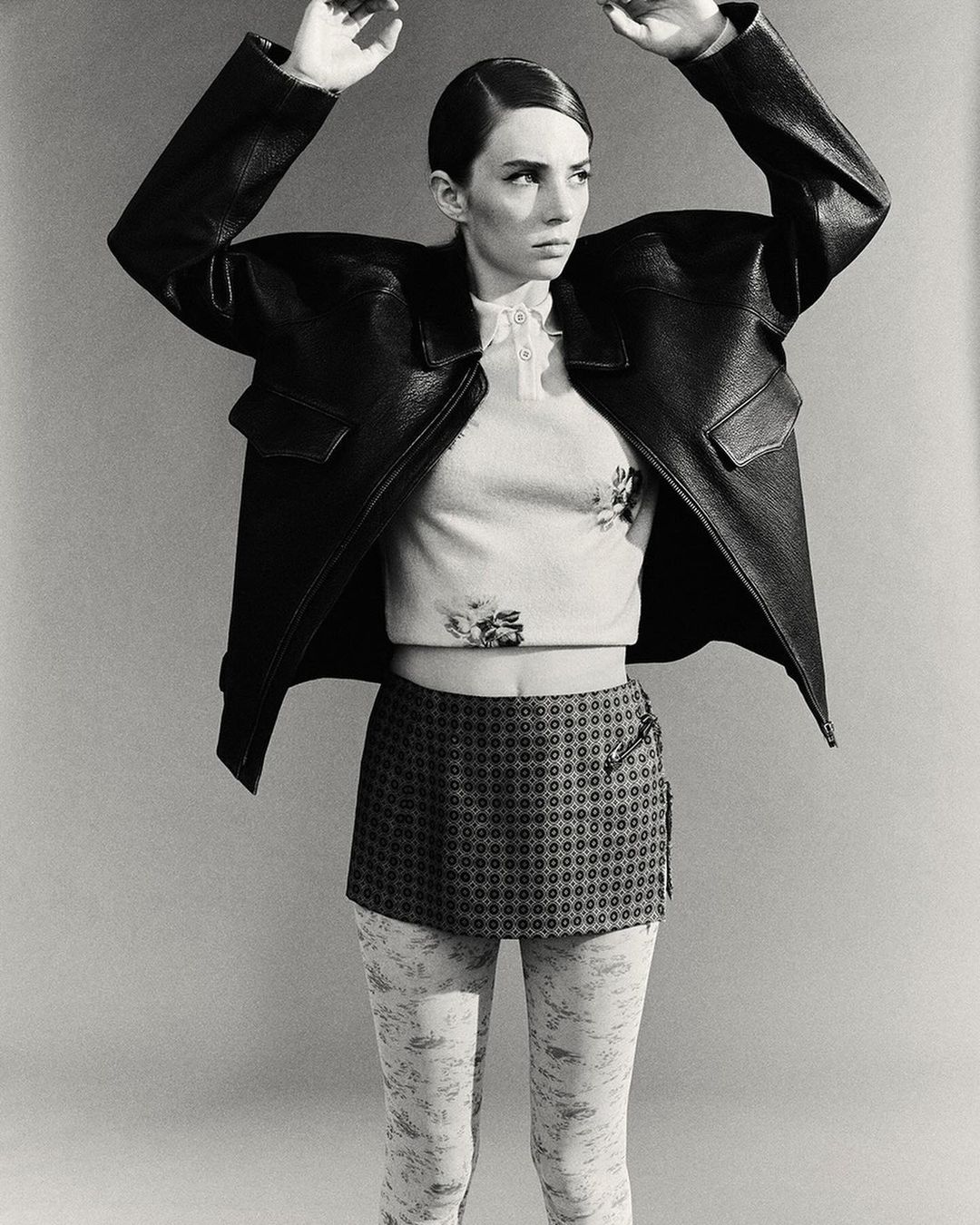Monochrome portrait of an adult female with hands up, wearing a black leather jacket, printed top, and patterned mini skirt against a plain background.