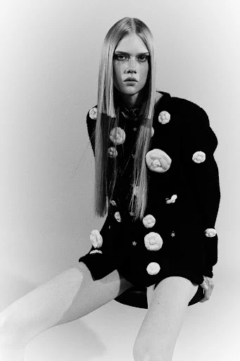 Young woman with fair skin and long blonde hair poses indoors wearing a black sweater dress with white rose appliqués in monochrome.