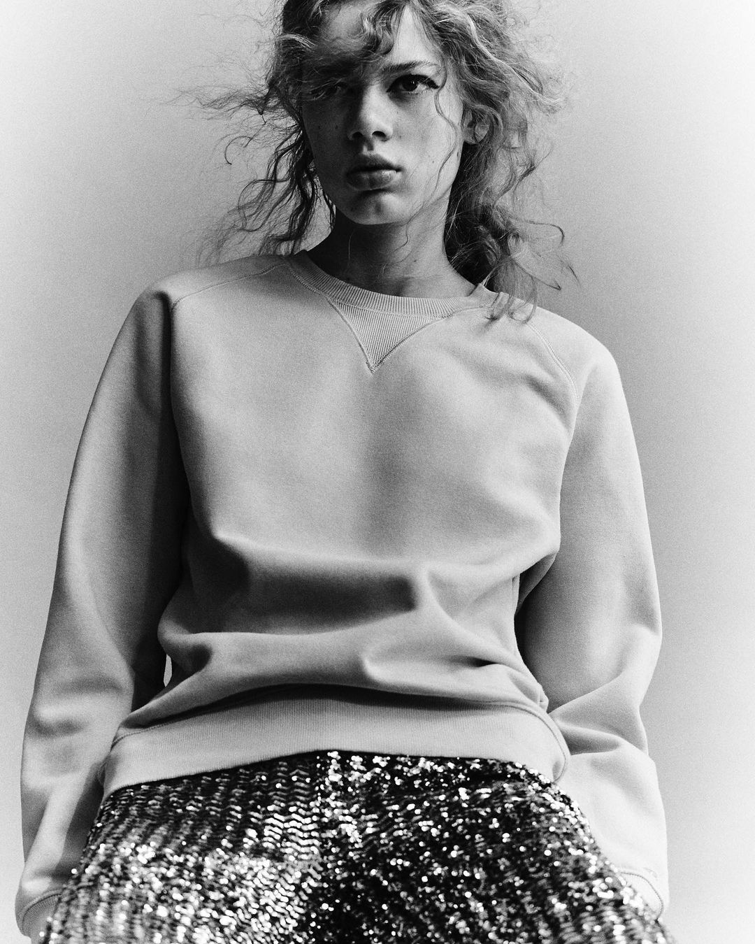 Monochrome fashion shot of a young adult female model in a crewneck sweatshirt and sequin pants looking directly at the camera.