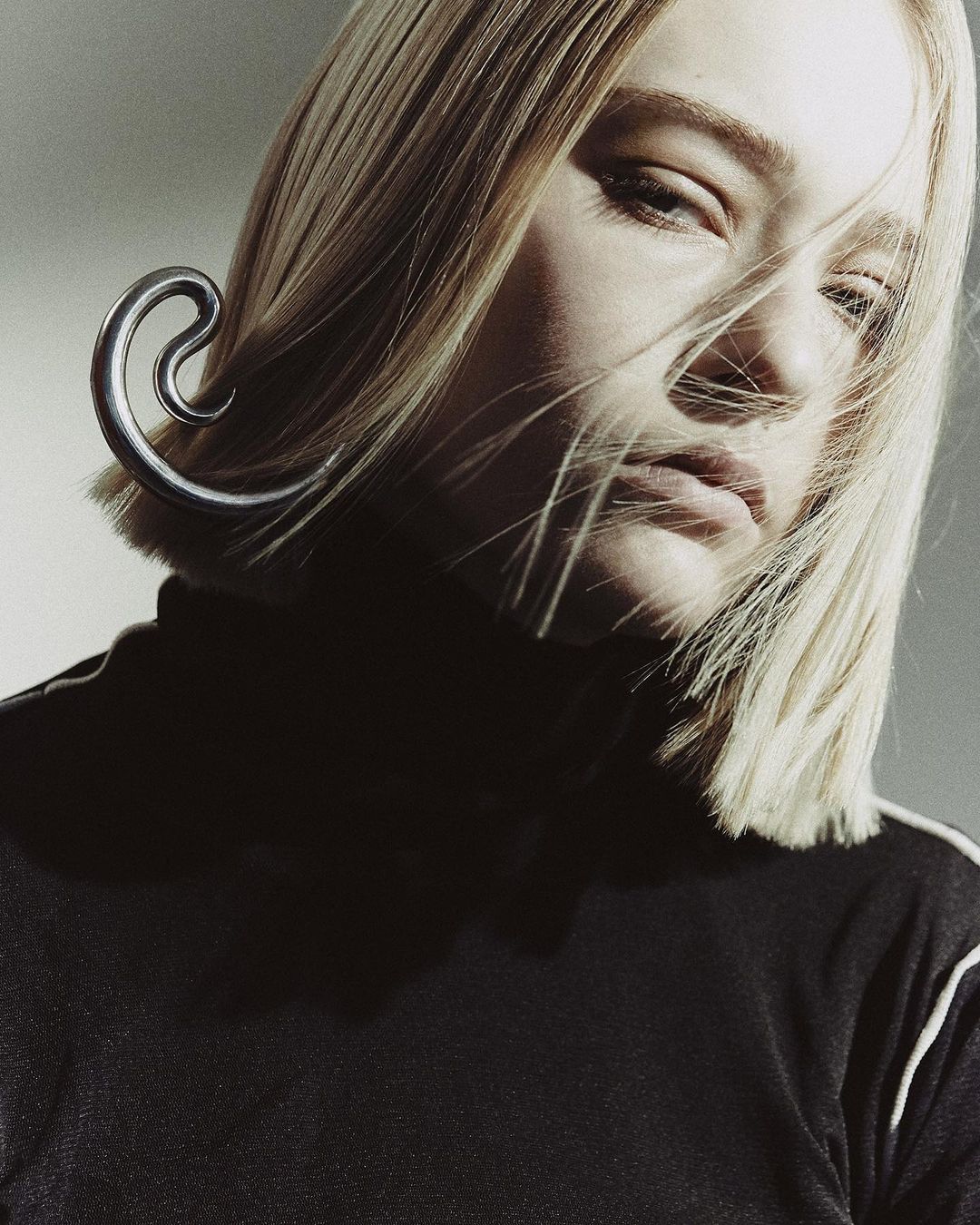 Close-up portrait of a blonde woman wearing a black turtleneck and a large metallic ear cuff by Melanie + Ramon