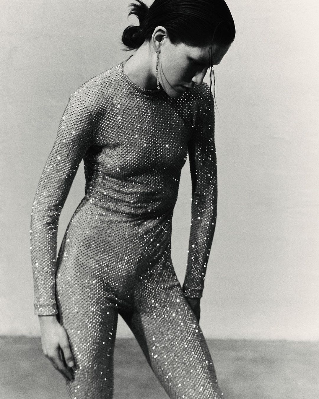 Black and white portrait of a woman in a sparkling, long-sleeved jumpsuit looking down, with dangling earrings and her hair pulled back.