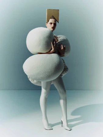 Model wearing an avant-garde padded white sculptural garment and cardboard headpiece against a grey studio backdrop.
