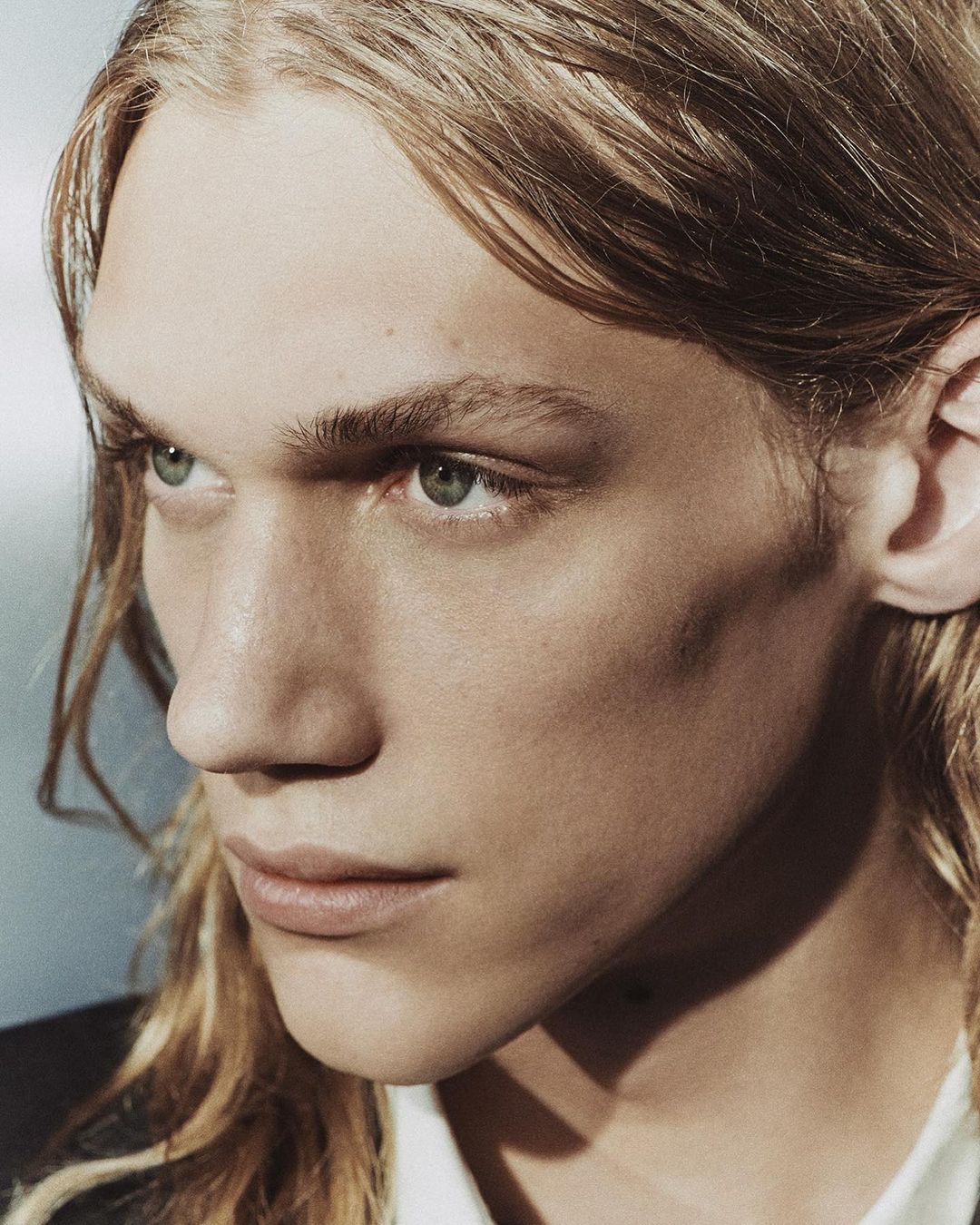 Portrait of a blonde male model with long hair, facing slightly left, featuring an eye-level angle in a studio setting.
