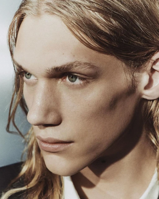 Portrait of a blonde male model with long hair, facing slightly left, featuring an eye-level angle in a studio setting.