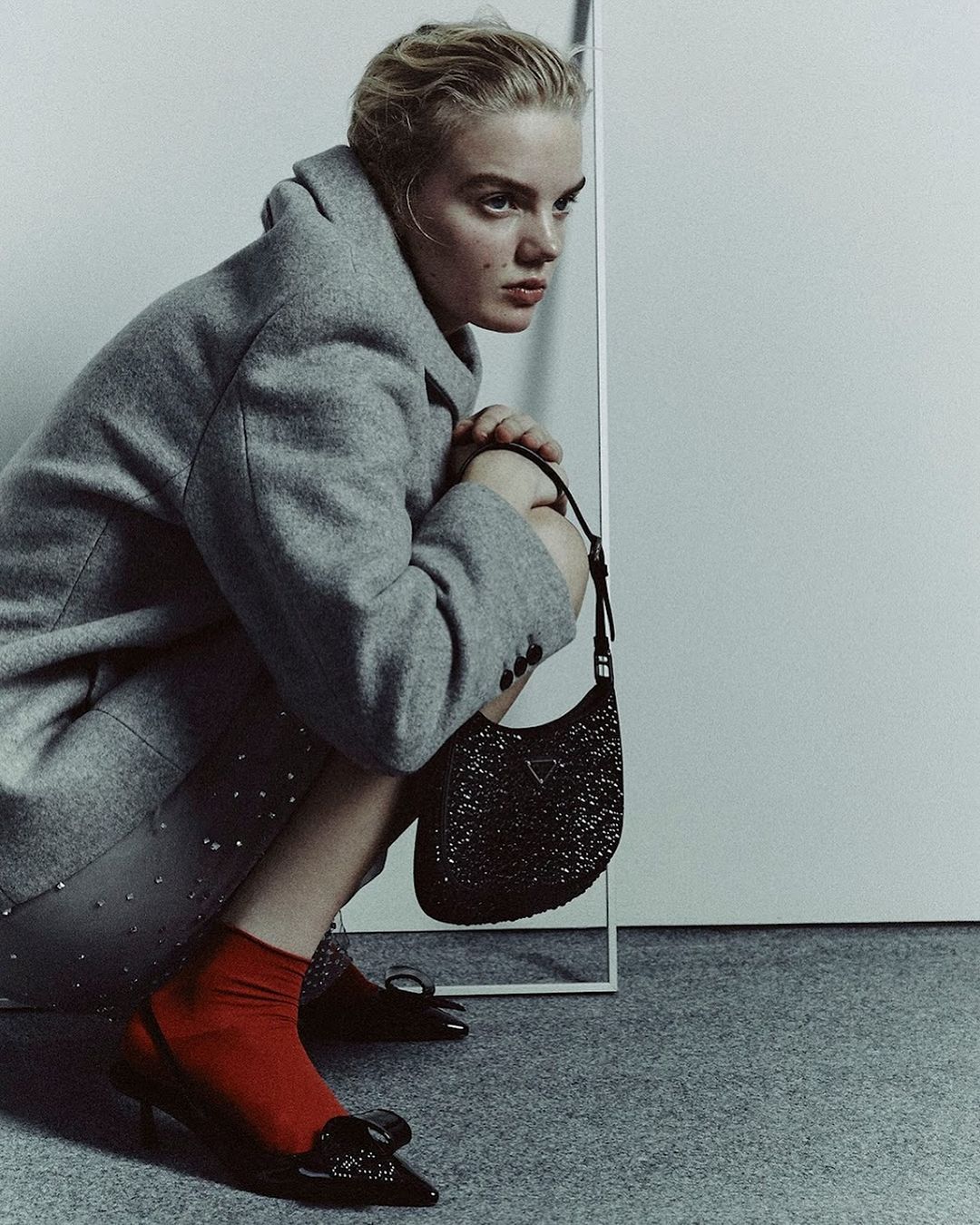 Young woman crouches in a grey coat, red socks, and black heels holding a black Prada purse, gazing away in an indoor setting.