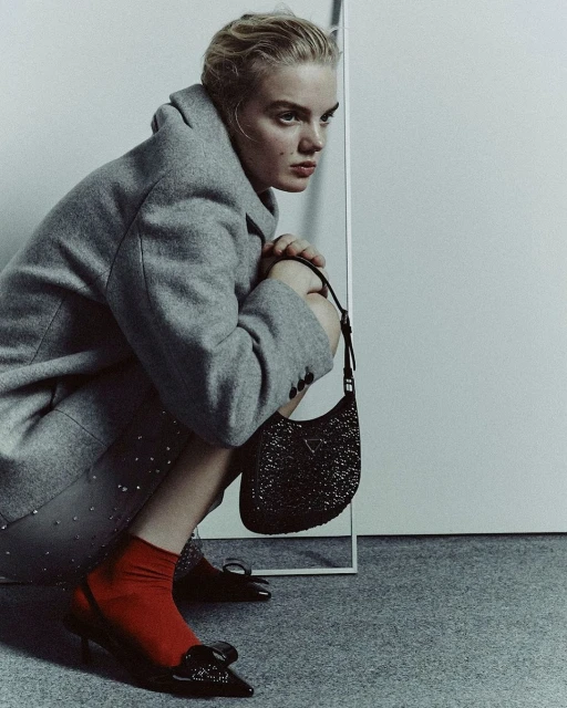 Young woman crouches in a grey coat, red socks, and black heels holding a black Prada purse, gazing away in an indoor setting.