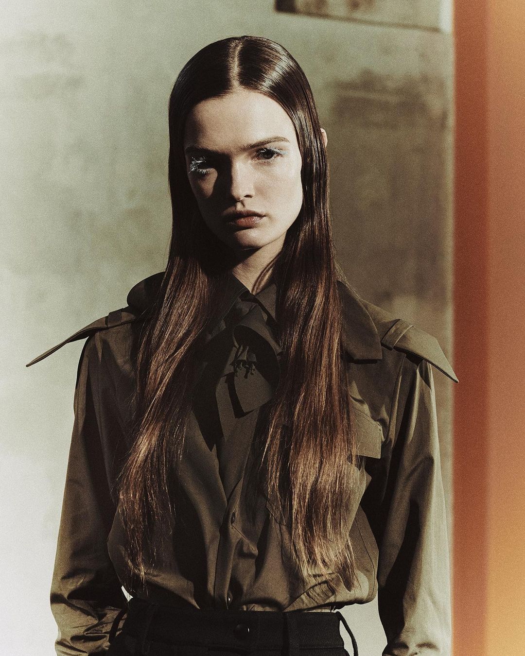 Portrait of a model with straight brown hair, wearing a dark green jacket, against a muted background with orange and grey sections.