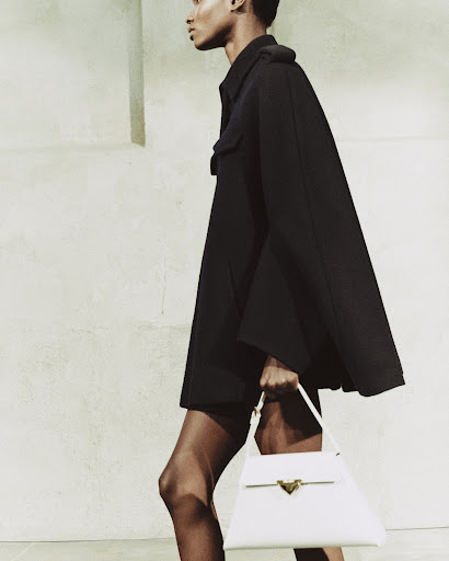 An adult female model walks in a fashion shot, wearing a black dress, and carrying a large, white leather handbag with a gold clasp.