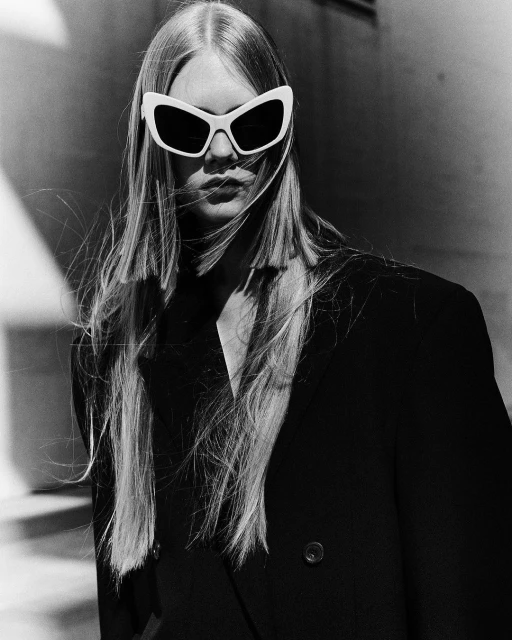 Monochrome portrait of a woman with long hair wearing sunglasses and a dark blazer against a neutral wall, black and white.