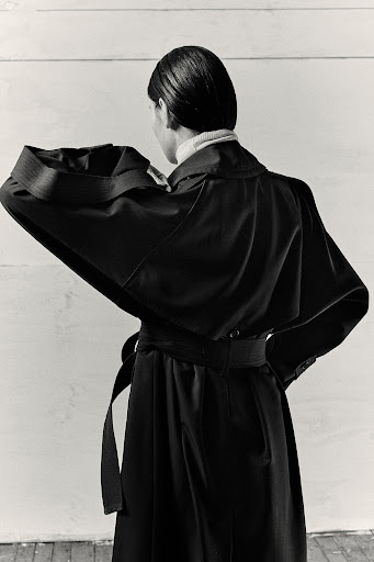Model seen from behind wearing an oversized dark belted coat against a white wooden paneled wall