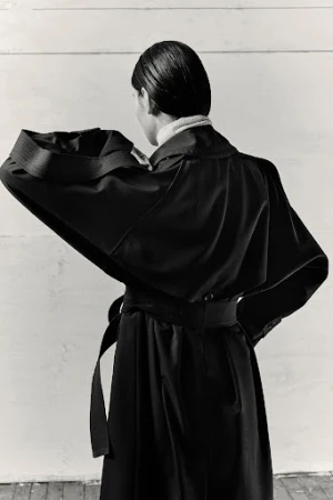 Model seen from behind wearing an oversized dark belted coat against a white wooden paneled wall