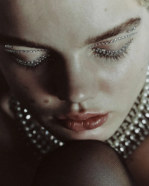 Close-up portrait of a woman with face jewelry, closed eyes, and glossy lips, set against a dark background, showcasing beauty and fashion.
