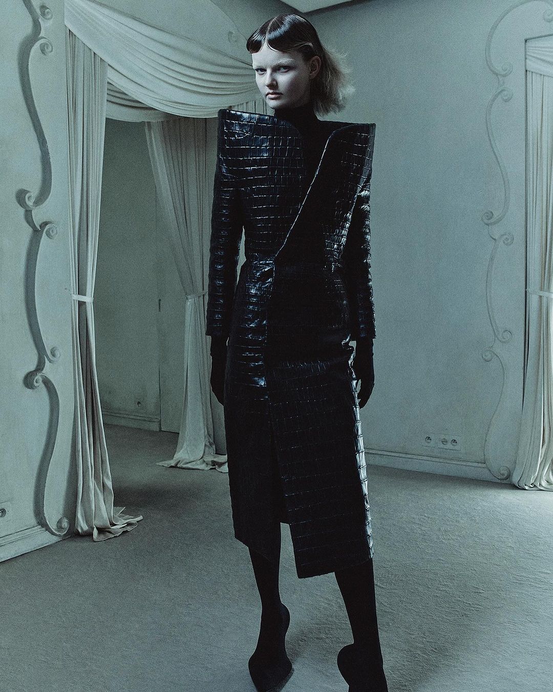 Elegant woman poses in a black crocodile-skin dress with strong shoulders in an ornate vintage interior with draped fabric and architectural moulding.