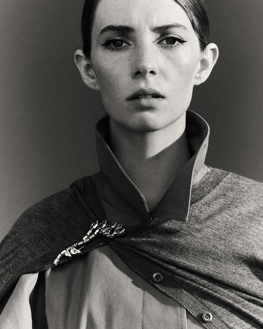 Portrait of a woman with dark eyeliner wearing a button-down shirt, grey cardigan, and jeweled brooch against a grey backdrop.