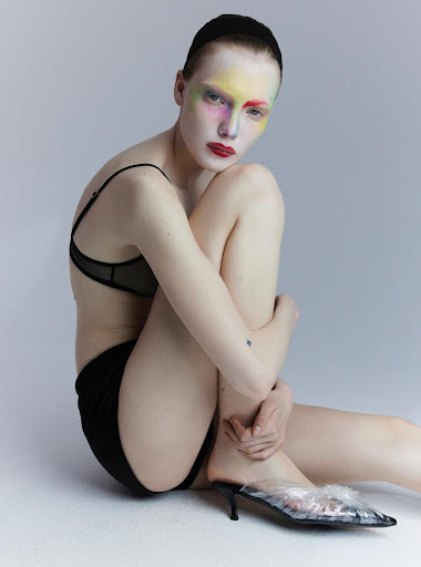 Woman poses in black lingerie and transparent heels, featuring rainbow makeup and red lipstick against a pale gray backdrop.