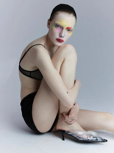 Woman poses in black lingerie and transparent heels, featuring rainbow makeup and red lipstick against a pale gray backdrop.