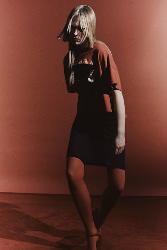 A blonde woman in a red top, black skirt, and red tights poses in front of a warm red background, fashion editorial photography.
