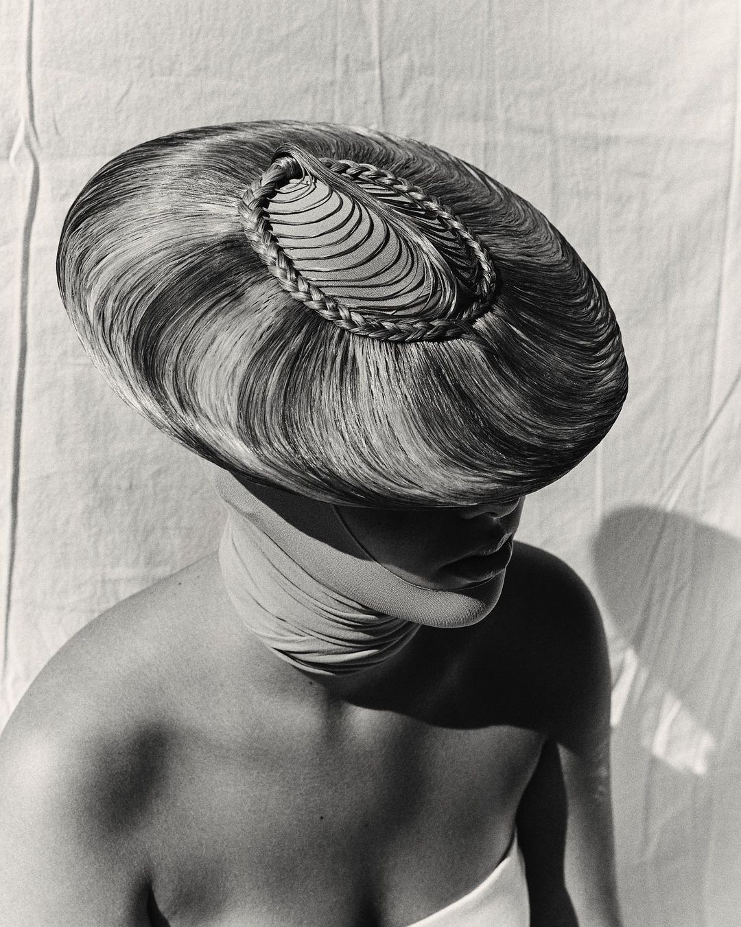 Striking monochrome portrait of a woman wearing an elaborate hairpiece and wrapped fabric, set against a textured off-white backdrop.