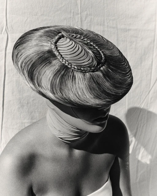Striking monochrome portrait of a woman wearing an elaborate hairpiece and wrapped fabric, set against a textured off-white backdrop.