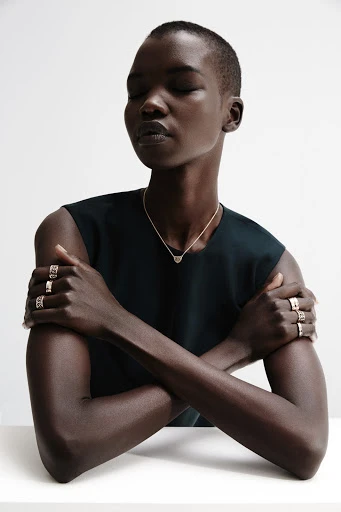Adult female with short dark hair wearing dark sleeveless shirt, silver necklace, and multiple rings posing indoors against a white wall.