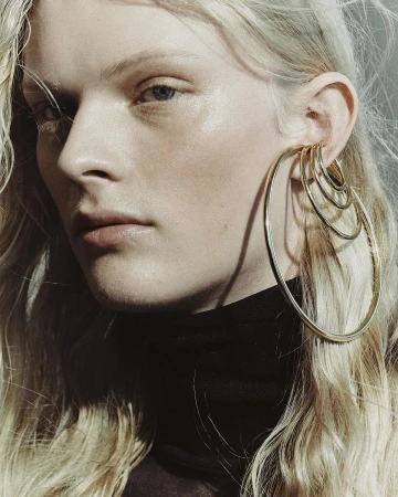 Close-up portrait of a model wearing stacked gold hoop earrings and a black turtleneck against a neutral background.