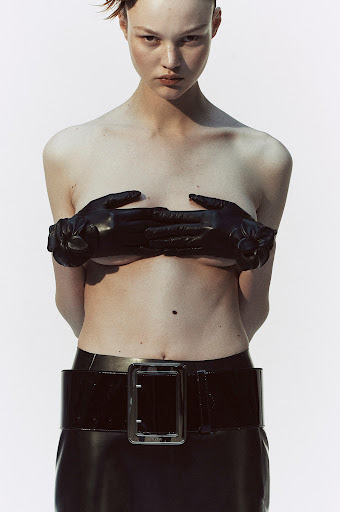 Fashionable woman with black leather gloves covering breasts, wears black patent leather skirt and belt against white backdrop in studio.