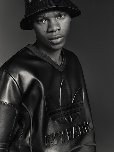 A young Black man wearing an Adidas x Ivy Park bucket hat and glossy, black, faux leather sweatshirt looks directly at the camera.