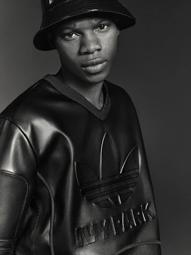 A young Black man wearing an Adidas x Ivy Park bucket hat and glossy, black, faux leather sweatshirt looks directly at the camera.