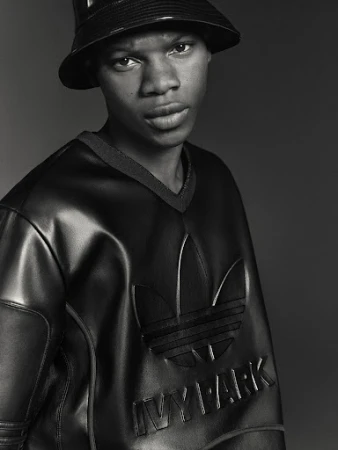 A young Black man wearing an Adidas x Ivy Park bucket hat and glossy, black, faux leather sweatshirt looks directly at the camera.