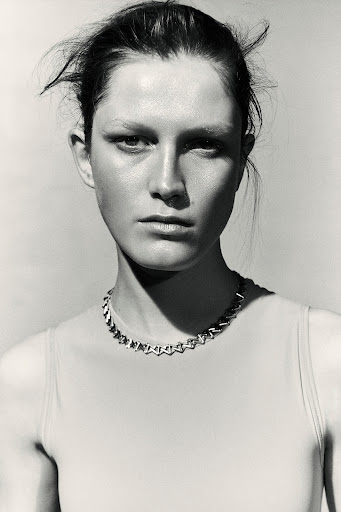 Close-up monochrome portrait of a woman wearing a geometric silver link necklace and sleeveless top against a light background.
