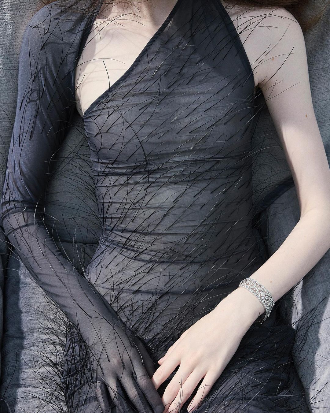 Adult woman in a fashionable black dress with one long sleeve. Bracelet on the wrist. Lying on grey fabric. Indoor fashion editorial.