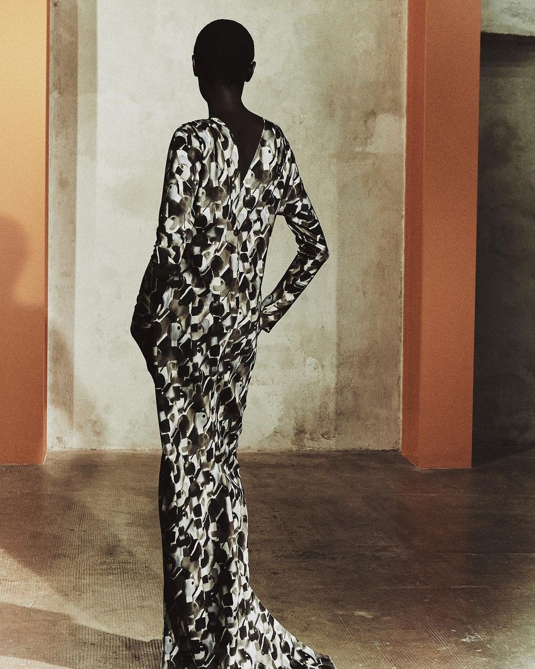 Woman poses indoors in a long-sleeved, patterned gown. She is facing away from the camera with an indoor concrete background.